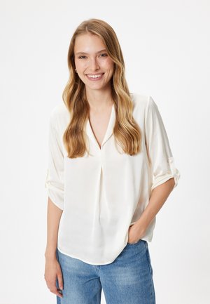 Helle Creme, seidige Bluse mit V-Ausschnitt und hochgerollten Ärmeln. Weiche Textur, lässiger Schnitt, ergänzt durch blaue Denim-Jeans.