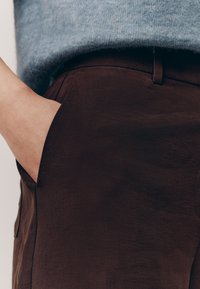 Pantaloni marroni con una texture liscia e una tasca laterale, abbinati a un maglione azzurro chiaro e soffice, che mostrano un contrasto di materiali e colori.