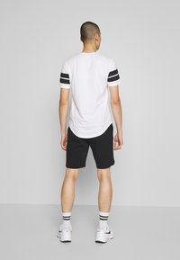 T-shirt blanche avec des rayures noires sur les manches courtes, associée à un short noir. Chaussures blanches avec des accents noirs et chaussettes blanches rayées.