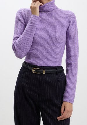 Femme portant un pull col roulé violet moulant en maille côtelée, rentré dans un pantalon noir à fines rayures, ceinturé avec une ceinture en cuir noire à boucle dorée.