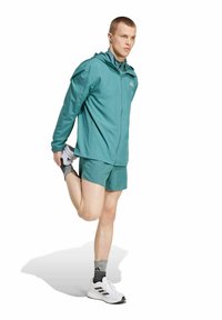 Chaqueta atlética color verde azulado y pantalones cortos, tejido ligero, cremallera frontal, modelo realizando un estiramiento. Zapatos blancos con detalles en negro, calcetines con estampado.