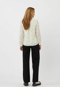 Hvid blondebluse med lange ærmer, blomsterdesign, indsnørede manchetter, parret med sorte bukser og lyse sko. Håret falder til skuldrene.
