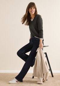 Ciemnozielony sweter, biała podkoszulka, ciemne jeansy z kontrastowym przeszyciem, trzyma beżową polarową kurtkę, białe buty z czerwonym akcentem.