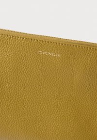 Mosterdgele leren etui met een zichtbare ritssluiting, een getextureerd oppervlak en het geperste "Coccinelle" logo in een modern lettertype.