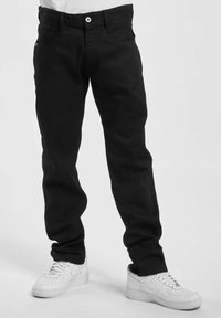 Bena iført sorte jeans og hvite Nike-sneakers, stående foran en ensfarget hvit bakgrunn.