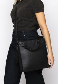 Sac à main en cuir noir avec une surface texturée, doté d'une bandoulière tressée et d'un accent à pompon. Forme rectangulaire avec un design structuré.