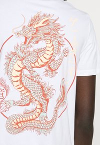 Bílý tričko s velkým ilustrovaným designem draka v oranžové a tyrkysové barvě na zadní straně, se složitými šupinami a vlnoucími detaily hřívy.