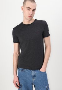 Schwarzes Kurzarm-T-Shirt mit Rundhalsausschnitt aus weichem Baumwollgemisch, mit einem kleinen gestickten Logo auf der Brust. Das Modell trägt eine blaue Jeans.