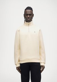 Pull beige clair avec col montant, demi-fermeture éclair et logo vert brodé. Le tissu semble doux avec une texture lisse et régulière.