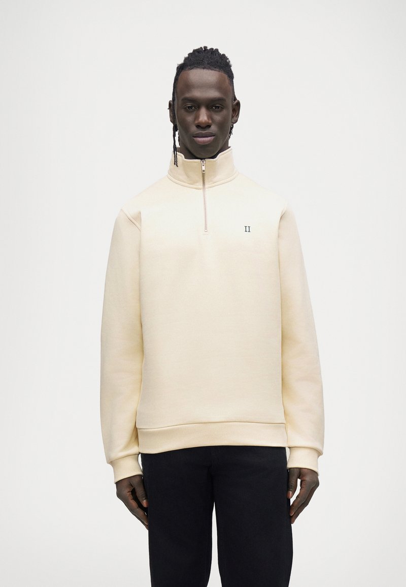 Pull beige clair avec col montant, demi-fermeture éclair et logo vert brodé. Le tissu semble doux avec une texture lisse et régulière.