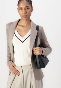 Béžový blazer přes krémový top s vlnitým výstřihem, spárovaný se světle šedými kalhotami. Drží malou černou kabelku se strukturovaným designem.