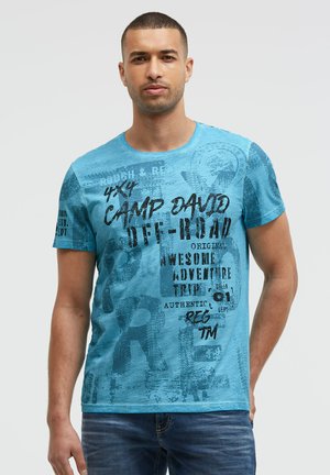 Man with short hair wearing a blue graphic t-shirt with black printed text and dark blue jeans standing against a plain light background.