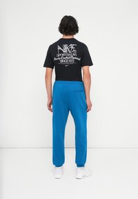 Μαύρο t-shirt με λευκό γραφικό τύπωμα στο πίσω μέρος και μπλε φόρμες. Οι φόρμες διαθέτουν πλαϊνές τσέπες, ελαστικούς σφιγκτήρες και μία πίσω τσέπη.