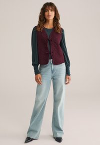 Gilet trapuntato borgogna con motivi floreali, annodato davanti; indossato sopra un top a maniche lunghe color teal e jeans larghi azzurri. Scarpe nere a punta.