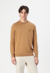 Pulover camel barve, narejen iz mehkega materiala, z okroglim izrezom, dolgimi rokavi in majhnim rumenim vezenim logotipom na prsih.