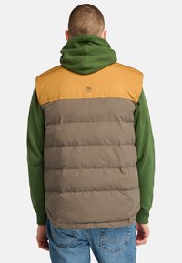 Puffy vest in brown and mustard yellow, with a zippered collar and a logo patch. Worn over a green hoodie, featuring a smooth texture.