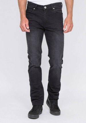Homme portant un jean noir slim-fit et des baskets noires, debout droit contre un fond clair uni.