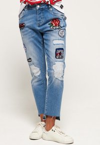 Blå denimjeans med en slitet design, som har olika färgglada lappar och broderier. Ankellängd med fransar.