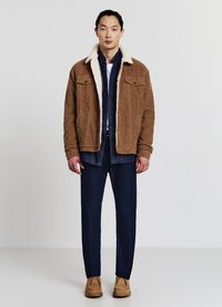 Braune Cordjacke mit einem cremefarbenen Kunstfellkragen, darunter ein navyfarbenes Hemd, dunkle blaue Jeans und hellbraune Schuhe. Einfaches, geschichtetes Design.