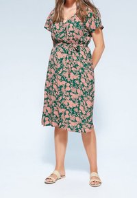 Vestido floral con escote en V, mangas cortas, fondo verde y patrón de flores rosas. Corte en A con cintura atada, combinado con sandalias beige.