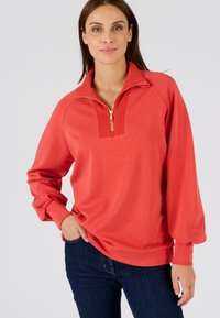 Koraalkleurige pullover met een kwart rits kraag, geribbelde details bij de hals en lange mouwen. Zacht materiaal met een losse fit. Draag het met een donkere spijkerbroek.