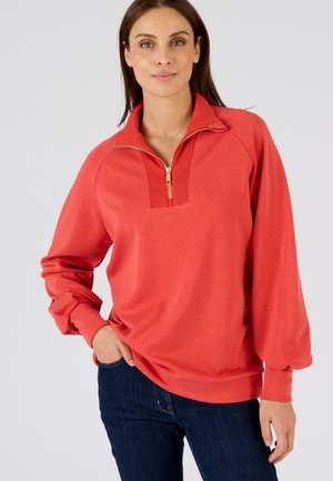 Koraalkleurige pullover met een kwart rits kraag, geribbelde details bij de hals en lange mouwen. Zacht materiaal met een losse fit. Draag het met een donkere spijkerbroek.