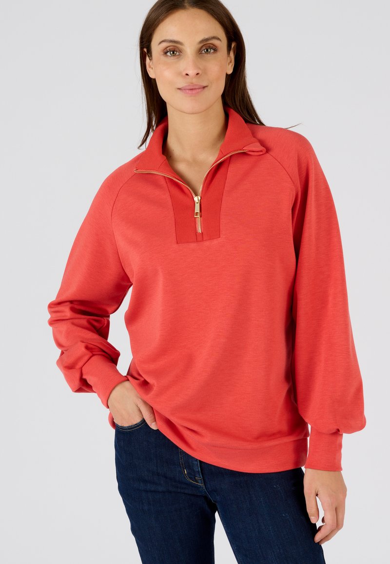 Pullover corallo con colletto a zip fino a un quarto, dettagli a coste sul collo e maniche lunghe. Tessuto morbido con una vestibilità rilassata. Indossato con jeans scuri.