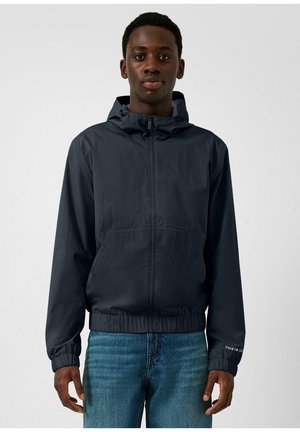 Jeune homme portant une veste à capuche zippée bleu marine et un jean bleu, debout devant un fond uni gris clair.