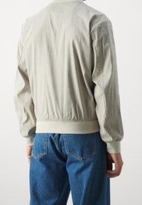 Světle béžová bomber bunda, bavlněný materiál, žebrované manžety a lem, elastický pas, viditelné švy, nošena s modrými džíny.