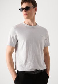 T-shirt en coton gris clair à manches courtes et col ras du cou. Présente un logo discret sur la poitrine. Associée à un pantalon noir et des lunettes de soleil.