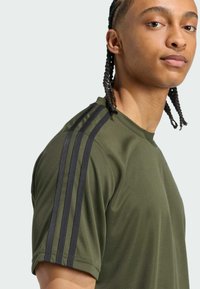Olivovo zelené športové tričko s krátkymi rukávmi, s tromi čiernymi pruhmi na pleciach a textúrovanou tkaninou vhodnou na šport.