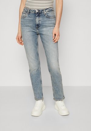 Person iført lyseblå jeans med lige ben, hvide sneakers og en sort-hvid stribet ærmeløs top, stående mod en ensfarvet baggrund.