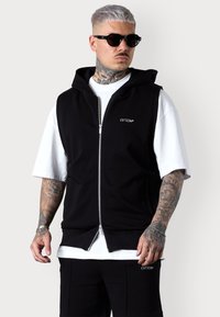 Schwarze ärmellose Kapuzenjacke mit einem Frontreißverschluss, ausgestattet mit einem weißen Logo auf der Brust und einem gerippten Saum. Getragen über einem weißen T-Shirt.