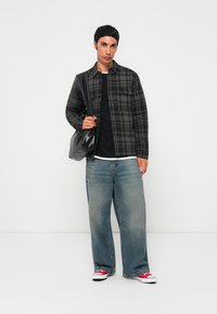 Young man standing wearing black and grey checkered jacket, black shirt, wide blue jeans, red sneakers, and carrying a black leather shoulder bag.