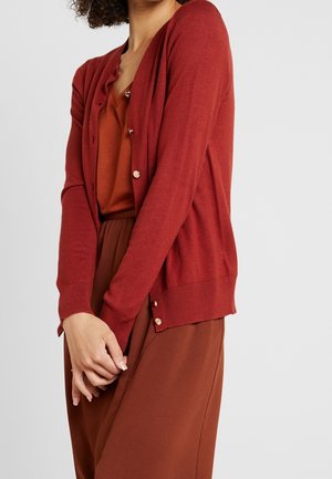 Kobieta w rdzawej marynarce na guziki, założonej na pasującą bluzkę i spódnicę w tym samym kolorze, stoi z delikatnie splecionymi rękami z przodu.