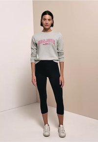 Femme debout portant un sweat gris avec le texte «BONJOUR», des leggings courts noirs et des baskets blanches sur un fond beige bicolore.