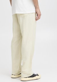 Lyse beige linnedragter med løs pasform, med en enkelt baglomme og opsmøgede kanter, kombineret med sorte sneakers og hvide snørebånd.