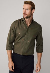 Man met lichtbruin haar en baard, gekleed in een olijfgroene button-up shirt met kleine rode florale patronen, met opgerolde mouwen en zwarte broeken.