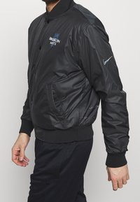 Chaqueta bomber negra con un acabado brillante, que presenta el logo de los Brooklyn Nets, botones a presión y puños elásticos. Incluye bolsillos laterales y detalles de Nike.