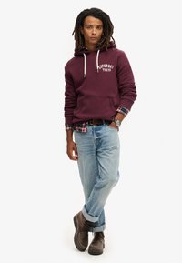 Felpa bordeaux con logo bianco, jeans blu strappati e stivali marroni con lacci. Presenta una camicia a quadri sotto e maniche arrotolate.