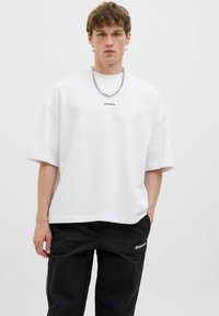 T-shirt ample blanc en coton doux avec des manches courtes, arborant un petit logo "CORE CREATIVE" et associé à un pantalon cargo noir.