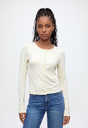 Vrouw met gevlochten haar, gekleed in een nauwsluitende crème lange mouwen top en blauwe jeans, staand tegen een effen lichte achtergrond.