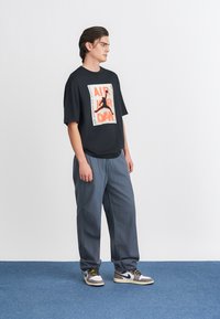 Μαύρο φανελάκι oversized με πορτοκαλί σιλουέτα, γκρι παντελόνι σε χαλαρή γραμμή και μαύρα και άσπρα αθλητικά παπούτσια με κίτρινες κορδόνες.