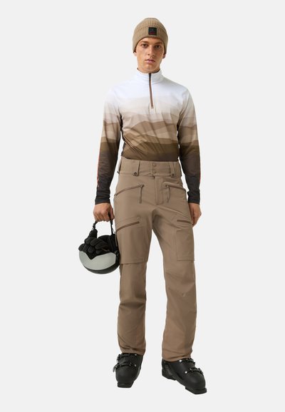 Skibekleidung bestehend aus einem langärmligen Oberteil mit weiß-braunem Farbverlauf, hellbraunen Hosen mit Taschen und schwarzen Skischuhen. Helm wird in der Hand gehalten.