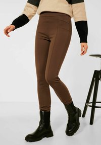 Bruna slim-fit byxor i stretchigt material, med strömlinjeformat design, sidofickor och en mjuk yta. Kombinerat med svarta stövlar.