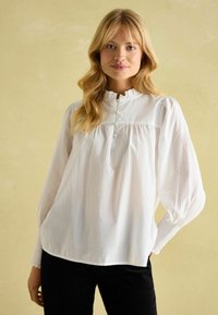 Weiße, langärmelige Bluse mit Rüschkragen, Knopfdetails und geraffter Taille. Der Stoff wirkt leicht und leicht strukturiert.
