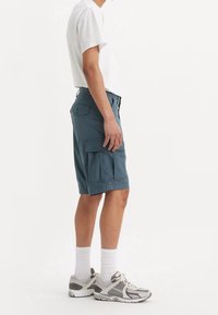 Osoba v bílém tričku, modrých cargo šortkách s několika kapsami, bílých kotníkových ponožkách a šedých a bílých sportovních botách, stojící bokem.