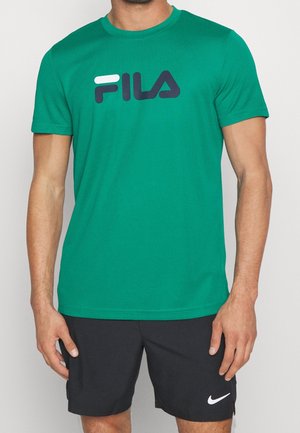 Uomo che indossa una maglietta verde FILA e pantaloncini neri Nike, in piedi contro uno sfondo chiaro e uniforme.