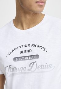 Hvit bomulls T-skjorte med rund hals. Har trykket tekst i svart og grått: "HEV DINE RETTIGHETER - BLEND BYGGT I BLÅ Vintage Denim."
