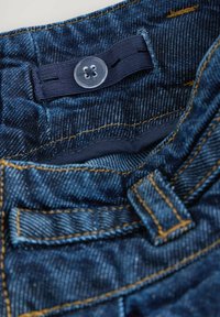 Jean en denim avec une teinte bleue, présentant des coutures orange, un bouton transparent et une ceinture texturée, mettant en avant plusieurs motifs de tissu.